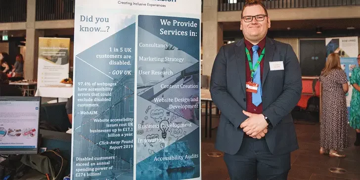 Image description: a business exhibition taking place at University of Staffordshire. Nick, a tall white cis male, is standing next to a promotional stand for Here4Inclusion, he has brown hair and is wearing glasses and a blue suit with a dark red shirt and light blue tie, he is looking directly at the camera and smiling. End of image description.