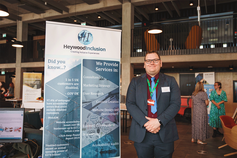 Image description: a business exhibition taking place at University of Staffordshire. Nick, a tall white cis male, is standing next to a promotional stand for Here4Inclusion, he has brown hair and is wearing glasses and a blue suit with a dark red shirt and light blue tie, he is looking directly at the camera and smiling. End of image description.