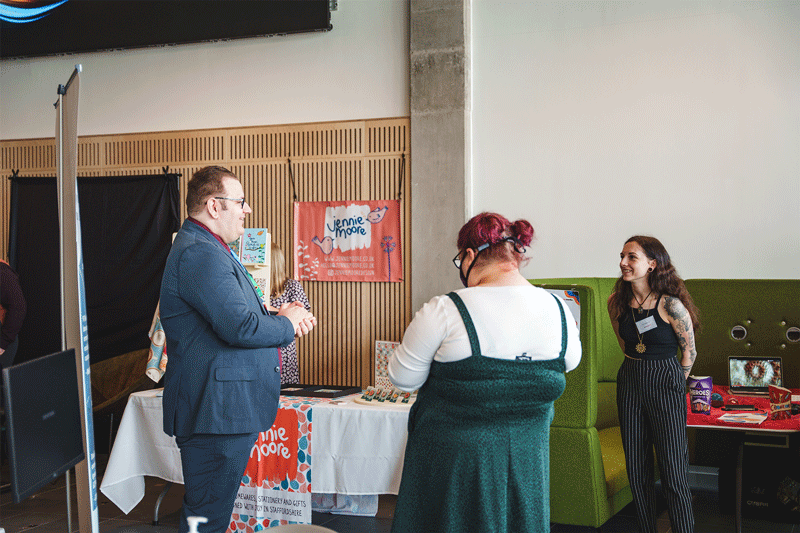 Image description: a business exhibition taking place at University of Staffordshire. Nick, a tall white cis male, is standing next to a promotional stand for Here4Inclusion, he has brown hair and is wearing glasses and a blue suit with a dark red shirt and light blue tie, he is speaking to two white cis women, while smiling, they are all looking at each other, unaware of the camera. End of image description.