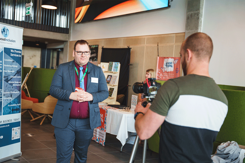 Image description: a business exhibition taking place at University of Staffordshire. Nick, a tall white cis male, is standing next to a promotional stand for Here4Inclusion, he has brown hair and is wearing glasses and a blue suit with a dark red shirt and light blue tie, he is looking away from the camera while taking part in an interview, facing another camera. End of image description.