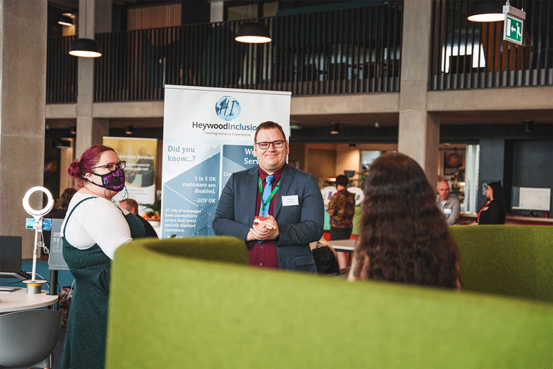 Image description: a business exhibition taking place at University of Staffordshire. Nick, a tall white cis male, is standing next to a promotional stand for Here4Inclusion, he has brown hair and is wearing glasses and a blue suit with a dark red shirt and light blue tie, he is speaking to two white cis women, while smiling, they are all looking at each other, unaware of the camera. End of image description.